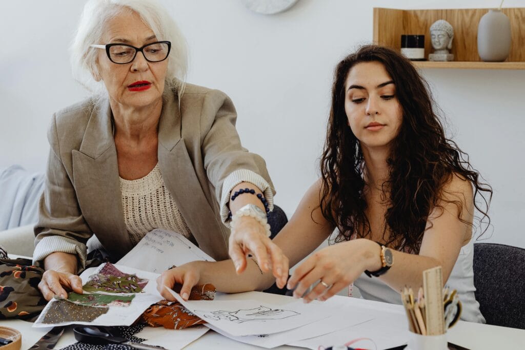 Older woman and younger woman sit next to each other at a table, reviewing drawings together. Photo by Kaboompics.com